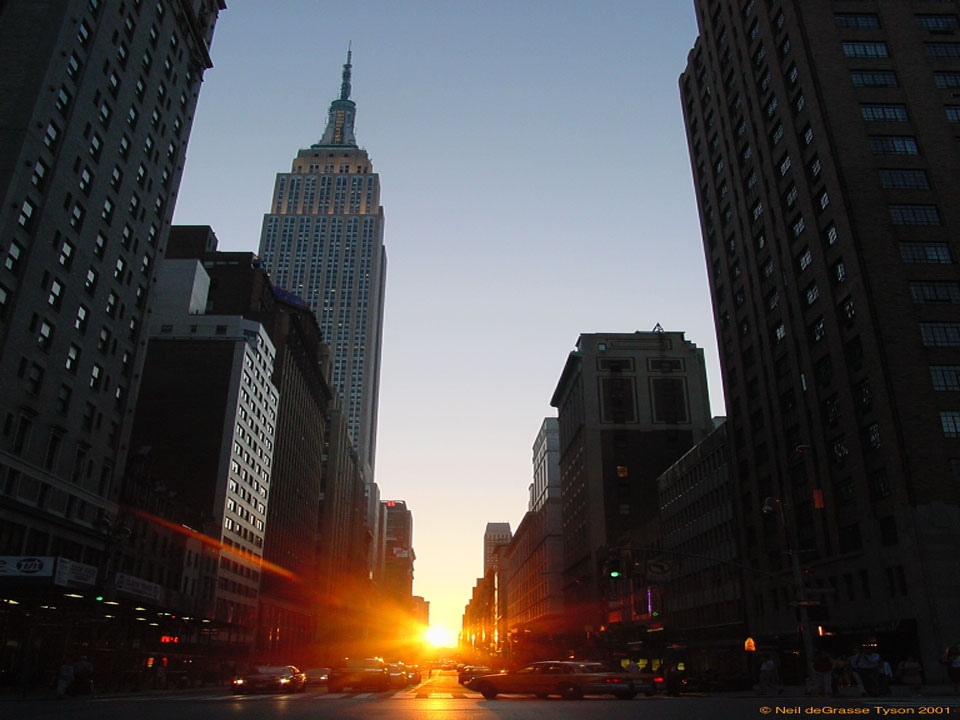 Die Sonne geht genau am Ende einer Häuserschlucht in Manhattan unter. Vorne fahren Autos, die Straße ist von Wolkenkratzern gesäumt, der Himmel blau und klar. Ein Klick auf das Bild zeigt das unverzerrte Bild.