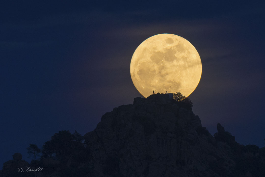 Der Erdmond steht hinter einem Felsenhügel. Der Mond ist fast voll. Auf dem Hügel ist die Silhouette einer Person zu sehen, die durch ein Teleskop blickt. Durch eine Überlagerung wird ein Teil des Mondes verdunkelt, der für manche wie ein Gesicht aussieht.