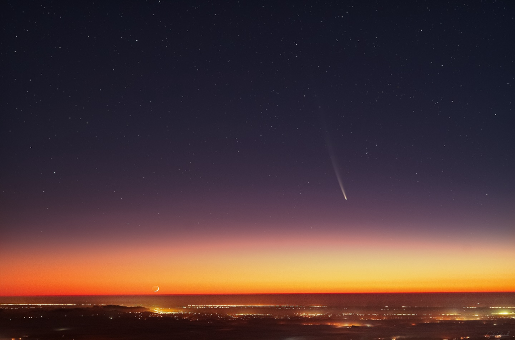 Über einer lichtverschmutzten Ebene leuchtet der Komet C/2023 A3 Tsuchinshan-ATLAS über dem Abendrot. Links am Horizont steht der schmale Sichelmond mit Erdlicht knapp über dem Horizont.